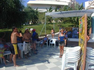 Mehrere Menschen versammeln sich im Freien auf einer Terrasse mit weißen Stühlen bei sonnigem Wetter.