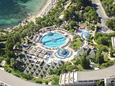 Luftaufnahme eines Resorts mit großen Pools, umgeben von grünen Bäumen und nahe am Meer gelegen.