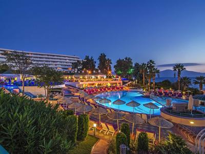 Hotelanlage mit beleuchtetem Pool, Palmen, grünen Pflanzen und blauem Abendhimmel am Meer.