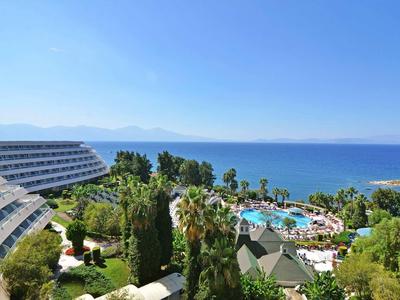 Hotelanlage mit Schwimmbad, grünen Bäumen und Meer im Hintergrund unter blauem Himmel.