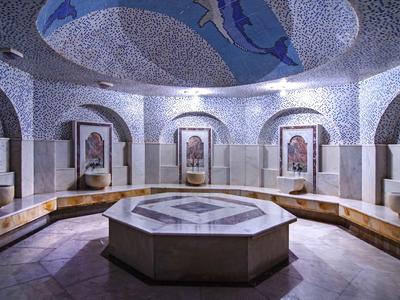 Runder Mosaikbrunnen in einem weißen, stilvollen Raum mit Mosaikboden und Sitzbank entlang der Wände.