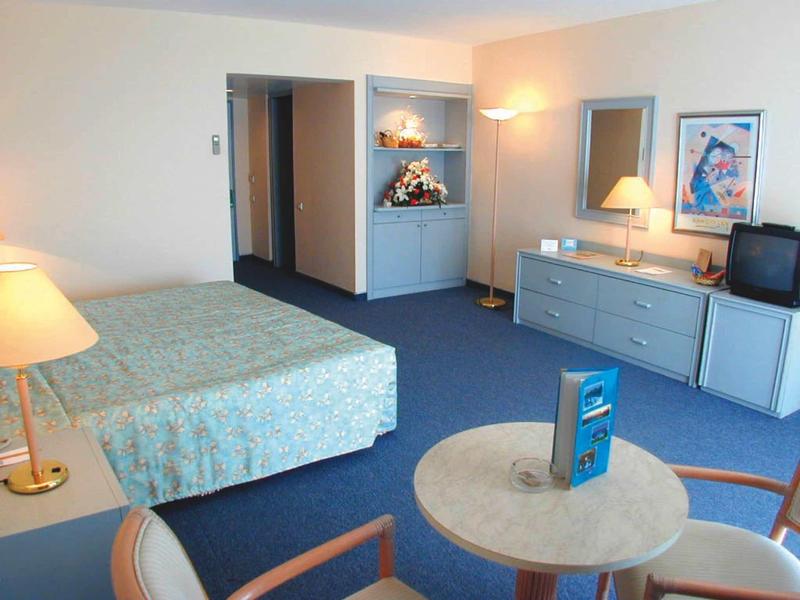 Hell eingerichtetes Hotelzimmer mit Bett, Schreibtisch, Stühlen und blauem Teppich.