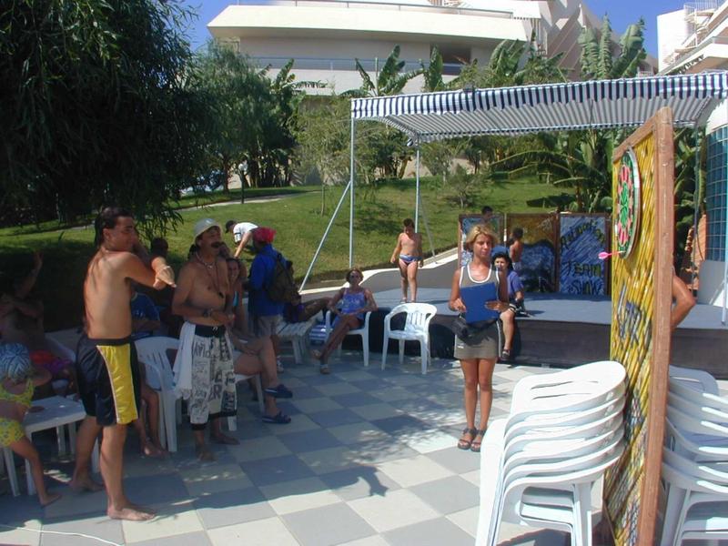 Menschen stehen in einem Kreis auf einer Terrasse, umgeben von Stühlen und grünen Bäumen.