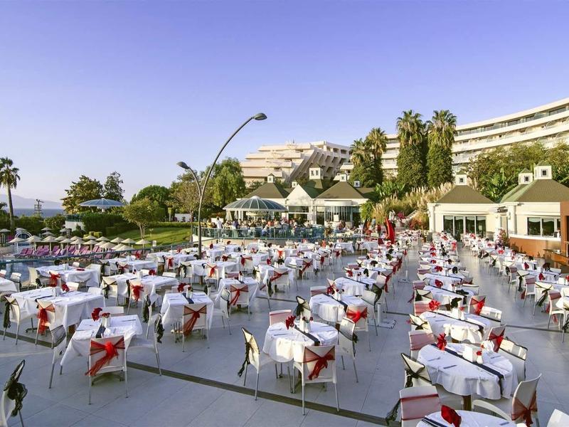 Großer Außenbereich mit vielen weiß gedeckten Tischen, Palmen und modernem Gebäude im Hintergrund.