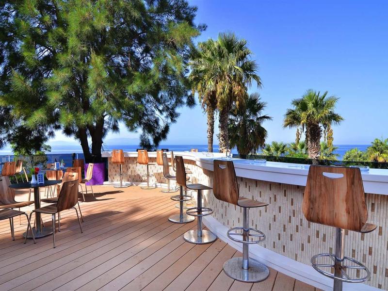 Freiluftbar mit Holzterrasse, Barhockern aus Holz, Tischen, Stühlen und Palmen im Hintergrund.