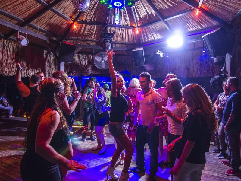 Menschen tanzen in einer Holzhütte mit strohgedecktem Dach und bunten Lichtern.