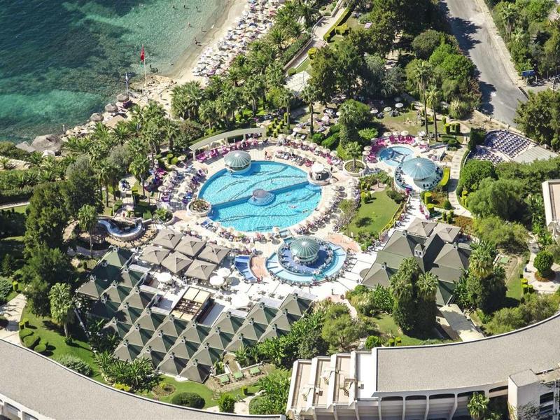 Luftaufnahme eines Resorts mit großen Pools, umgeben von grünen Bäumen und nahe am Meer gelegen.
