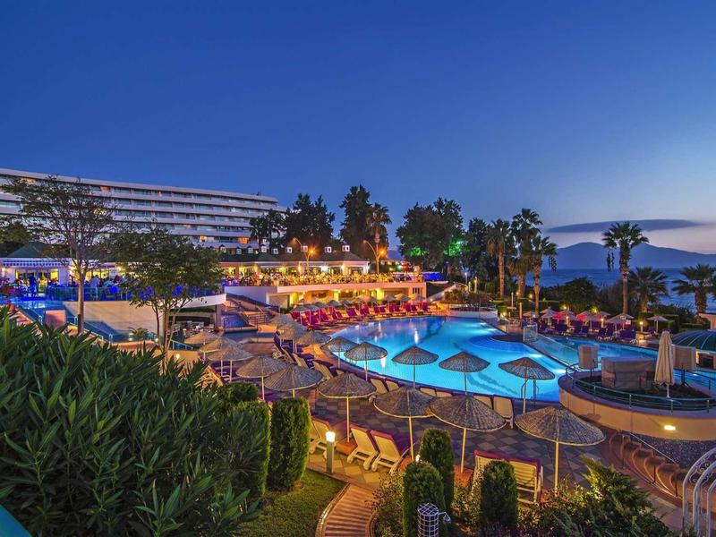 Hotelanlage mit beleuchtetem Pool, Palmen, grünen Pflanzen und blauem Abendhimmel am Meer.