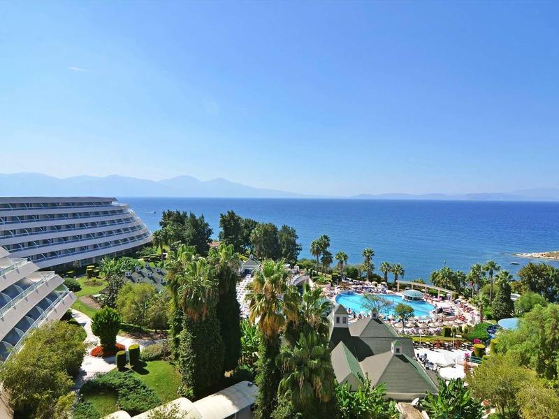 Hotelanlage mit Schwimmbad, grünen Bäumen und Meer im Hintergrund unter blauem Himmel.