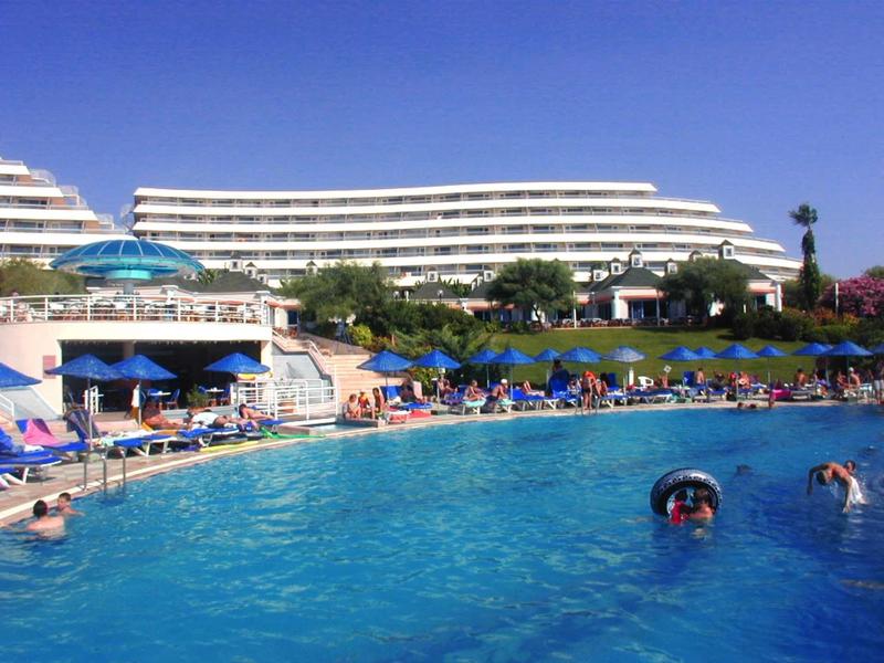 Großes Schwimmbecken mit Menschen, Sonnenschirmen und weißem Hotelgebäude im Hintergrund.