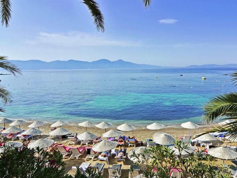 Strand mit Sonnenschirmen, Palmen und klarem türkisblauem Meer vor Bergen am Horizont.
