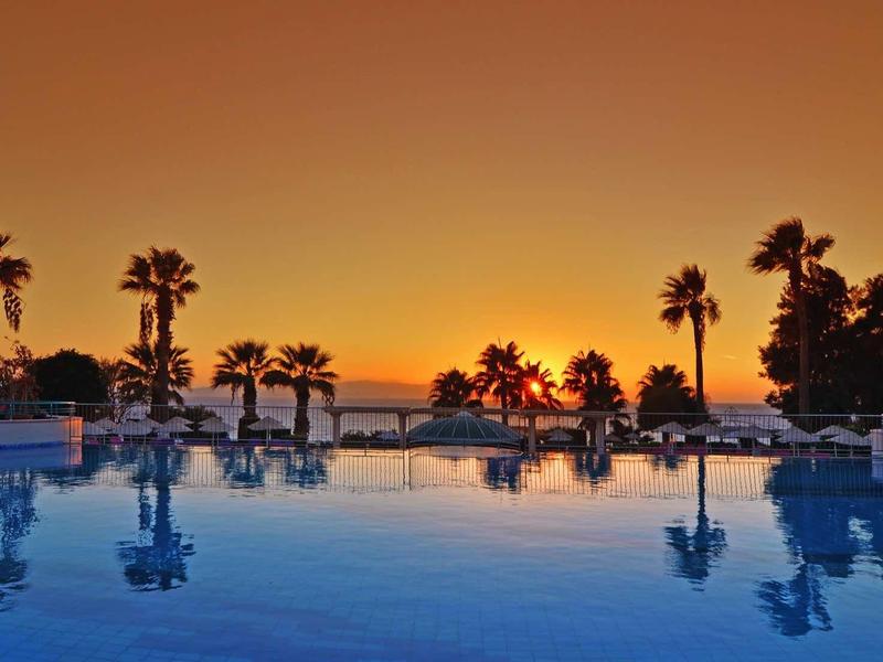 Blick auf einen ruhigen Pool mit Palmen, darunter ein Sonnenuntergang am Himmel.