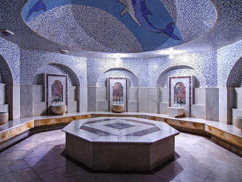 Runder Mosaikbrunnen in einem weißen, stilvollen Raum mit Mosaikboden und Sitzbank entlang der Wände.