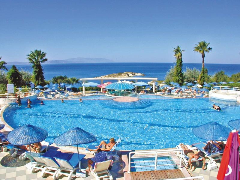 Großer Pool mit blauen Sonnenschirmen, Liegestühlen und Palmen vor klarem Meer und blauem Himmel.