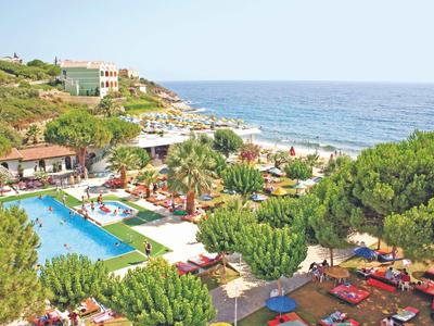 Blick auf ein Hotel mit großem Pool, grünen Bäumen und Blick aufs blaue Meer am bewölkten Himmel.
