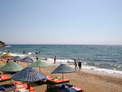Strand mit blauem Meer, sonnigem Himmel, vielen Sonnenschirmen und wenigen Menschen am Wasser.