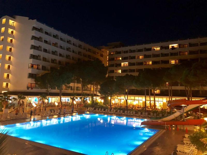 Beleuchteter Pool vor mehrstöckigem Hotelgebäude mit zahlreichen Fenstern und Palmen bei Nacht.