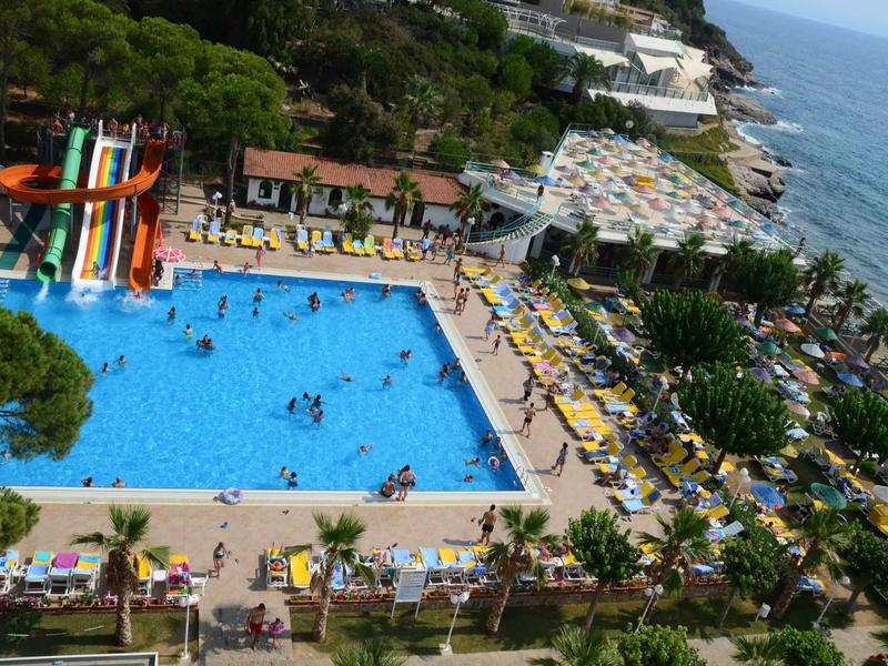 Großer blauer Pool mit bunten Wasserrutschen, Sonnenliegen und Palmen nahe einem Meer und Hügel.