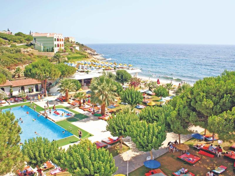 Blick auf ein Hotel mit großem Pool, grünen Bäumen und Blick aufs blaue Meer am bewölkten Himmel.
