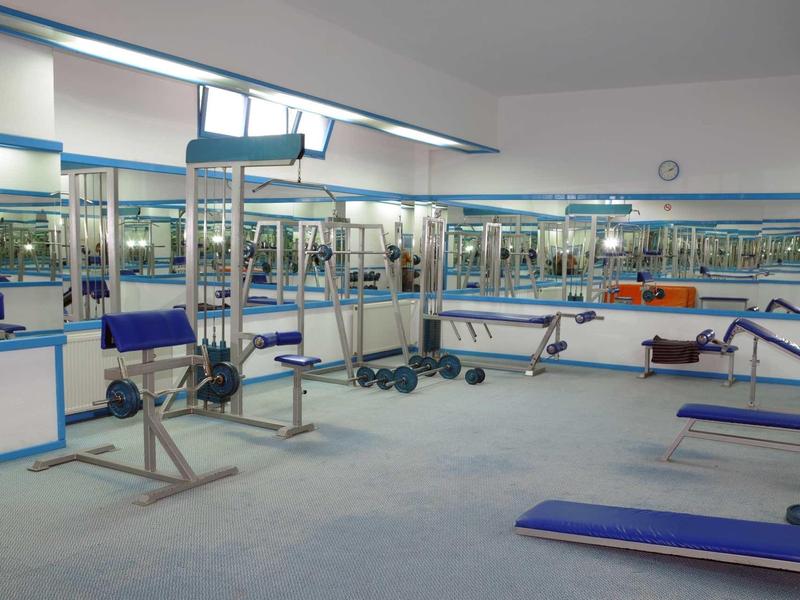 Weißer Fitnessraum mit blauen Trainingsbänken, Gewichten und großen Spiegeln an der Wand.