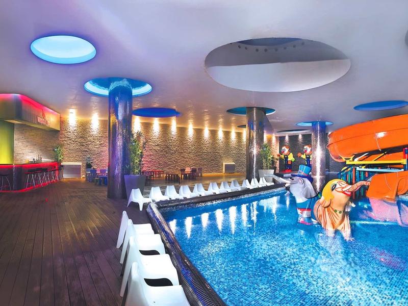 Innenraum eines modernen Hallenbads mit Wasserrutsche, Pool, bunten Lichtern und Holzverkleidung.