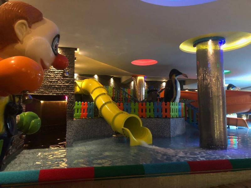 Bunter Indoor-Spielplatz mit gelber Rutsche, Wasserbecken und Clown-Figur an der Seite.