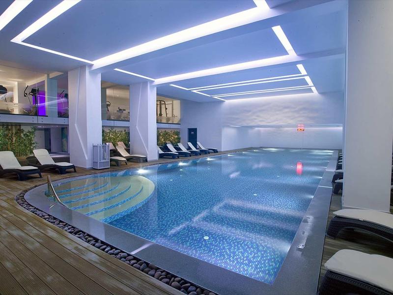 Moderner Innenpool mit blau leuchtendem Wasser, Liegestühlen und weißer Deckenbeleuchtung.