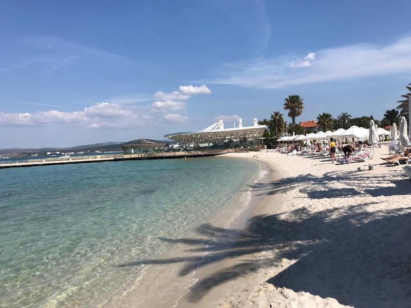 Strand mit klarem Wasser, weißem Sand und Palmen unter blauem Himmel und vereinzelten Wolken.