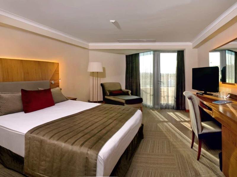 Schlafzimmer mit großem Bett, Holzdekor, Schreibtisch, Stuhl, Fernseher und Balkon mit Vorhängen.