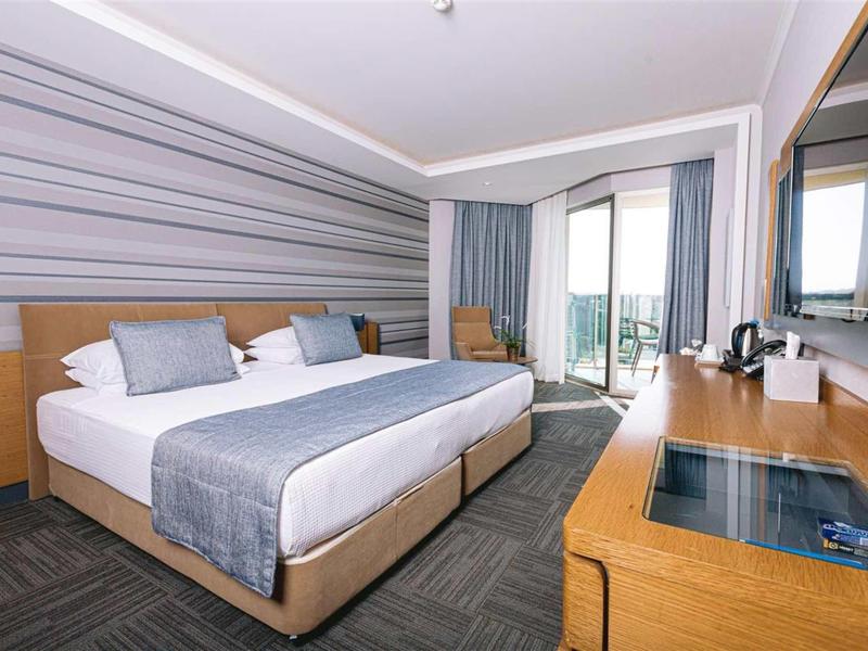 Modernes Hotelzimmer mit Doppelbett, Schreibtisch, Fernseher und Balkon mit Glasgeländer.