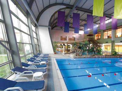 Hallenbad mit Schwimmbecken, blauen Liegestühlen, großen Fenstern und bunten hängenden Fahnen.