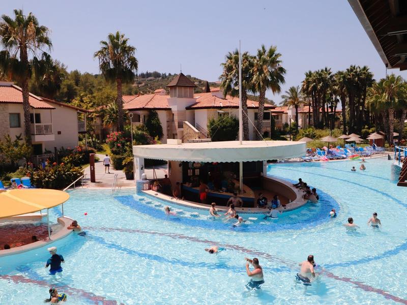 Großer Pool mit Bar in der Mitte, umgeben von Palmen und Häusern, viele Menschen im Wasser.