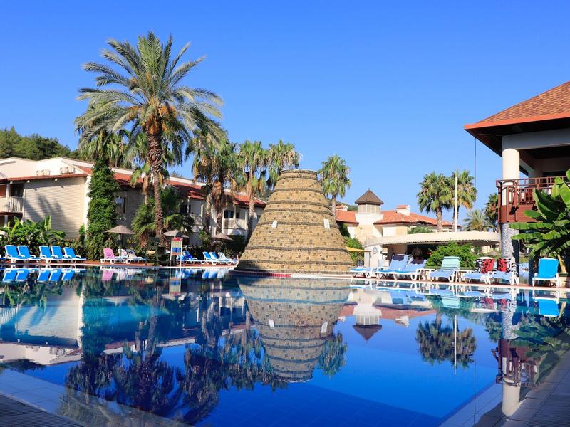 Blauer Pool mit Sonnenschirmen, Palmen und Gebäuden unter klarem Himmel in Tropenanlage.