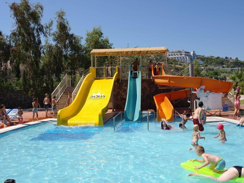 Wasserpark mit Rutschen in Gelb, Blau und Orange, Menschen spielen im blauen Pool unter blauem Himmel.
