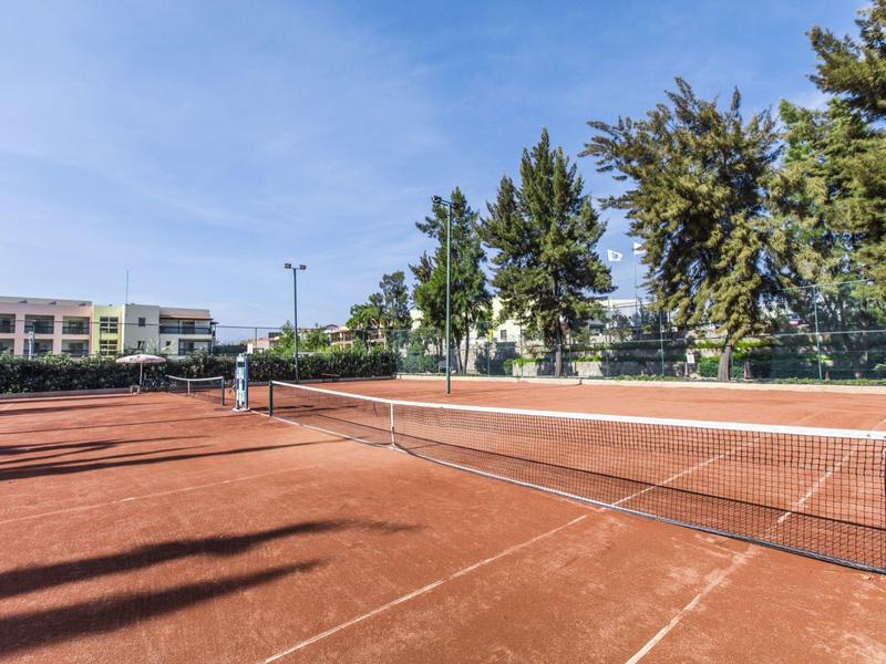 Roter Sand-Tennisplatz mit Netz, umgeben von Bäumen und Gebäuden unter blauem Himmel.