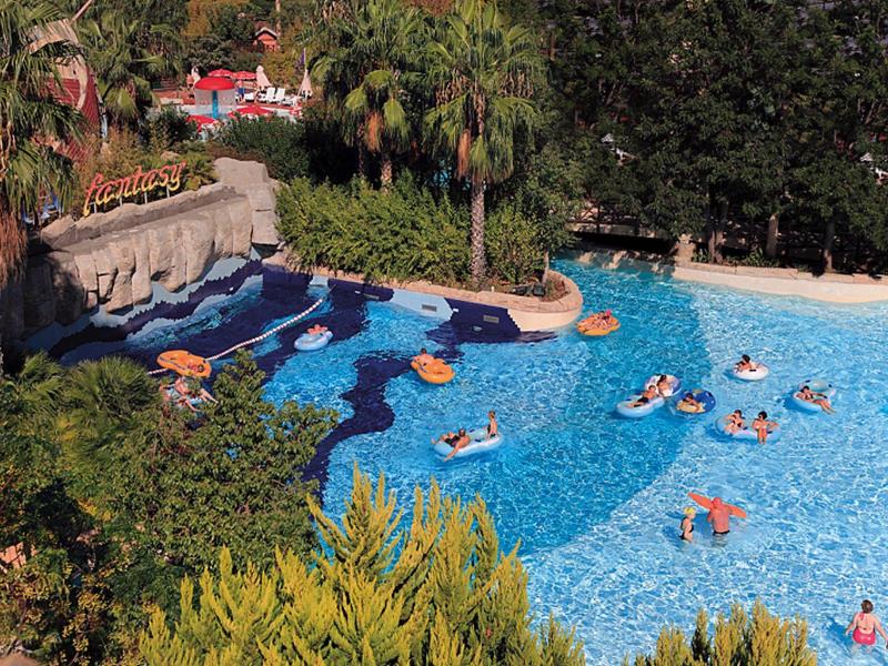 Großer Freizeitpool mit Personen auf Luftmatratzen, umgeben von Palmen und grünen Büschen.