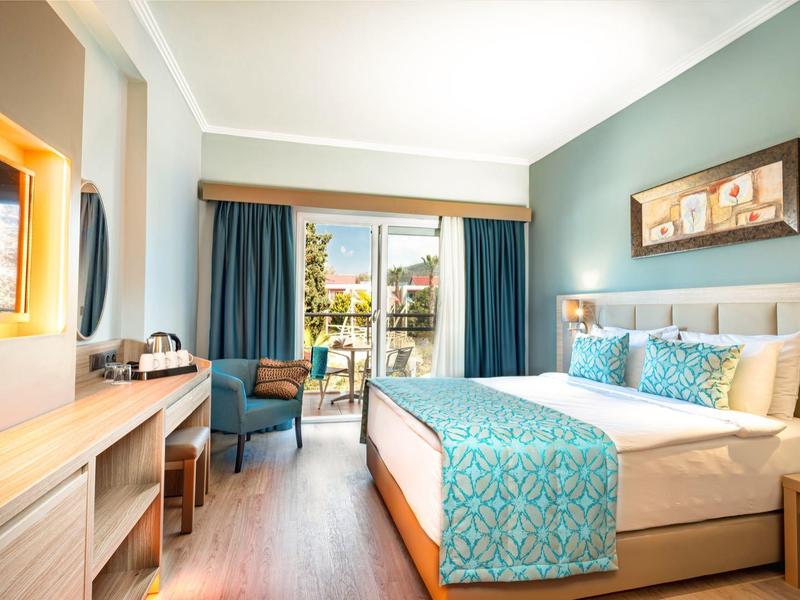 Helles Hotelzimmer mit großem Bett, blauem Bettdekor, Schreibtisch und Balkon mit Blick ins Grüne.