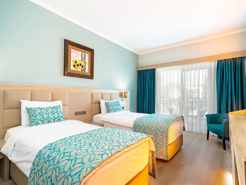 Heller Hotelzimmer mit zwei Einzelbetten, türkis gemusterten Decken, Holzfußboden und großen Fenstern.