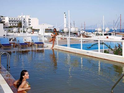 Zwei Frauen entspannen im Pool mit Blick auf Boote und weiße Gebäude am Meer bei klarem Himmel.