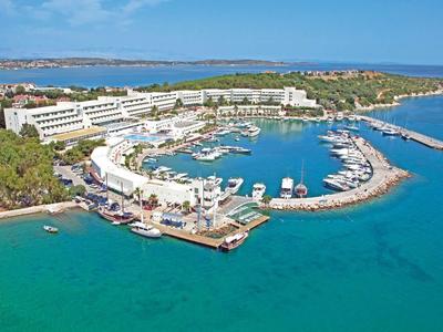 Blick auf einen Hafen mit vielen Booten, weißen Gebäuden und klarem blauem Wasser bei sonnigem Himmel.