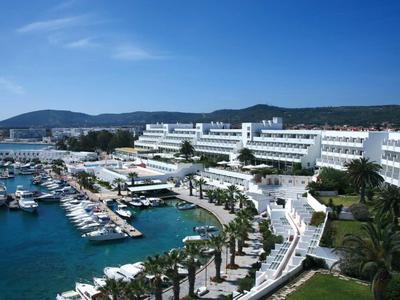 Großes weißes Hotel am Meer mit Yachthafen, Palmen und Bergen im Hintergrund unter blauem Himmel.