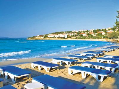 Strand mit vielen blauen Liegen, Sand, türkisfarbenem Meer und Häusern am Hügel unter blauem Himmel.