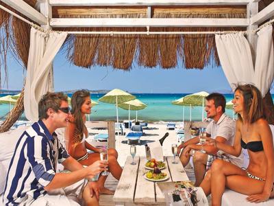 Strandbar mit Holzstruktur, Strohdach, weißen Vorhängen, sechs Menschen und Blick aufs Meer.