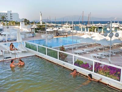 Hotelpool mit Gästen, Blick auf Liegestühle und Hafen im Hintergrund bei Sonnenlicht.