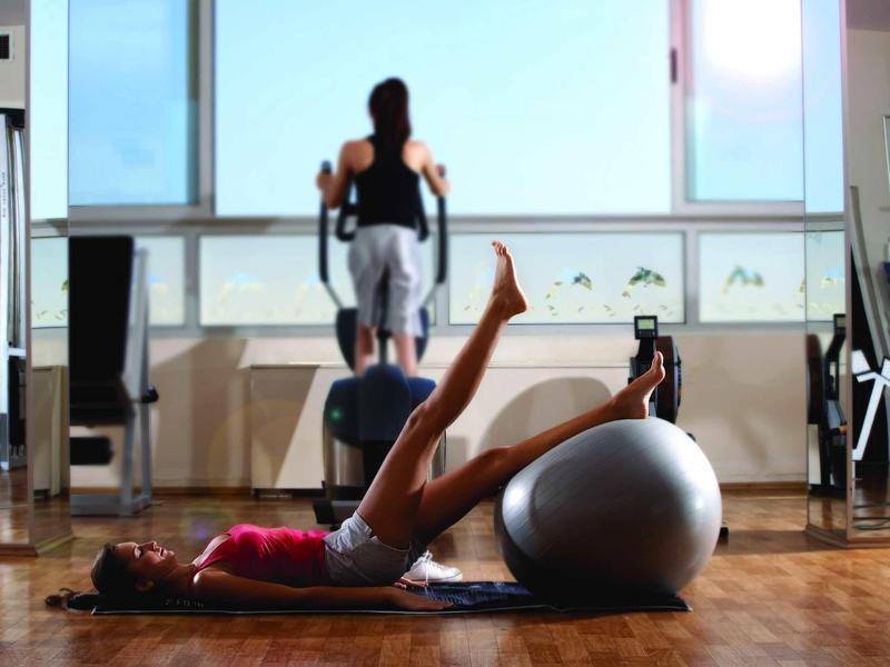 Frau trainiert mit Gymnastikball auf Boden, Mann läuft auf Laufband im Fitnessstudio.