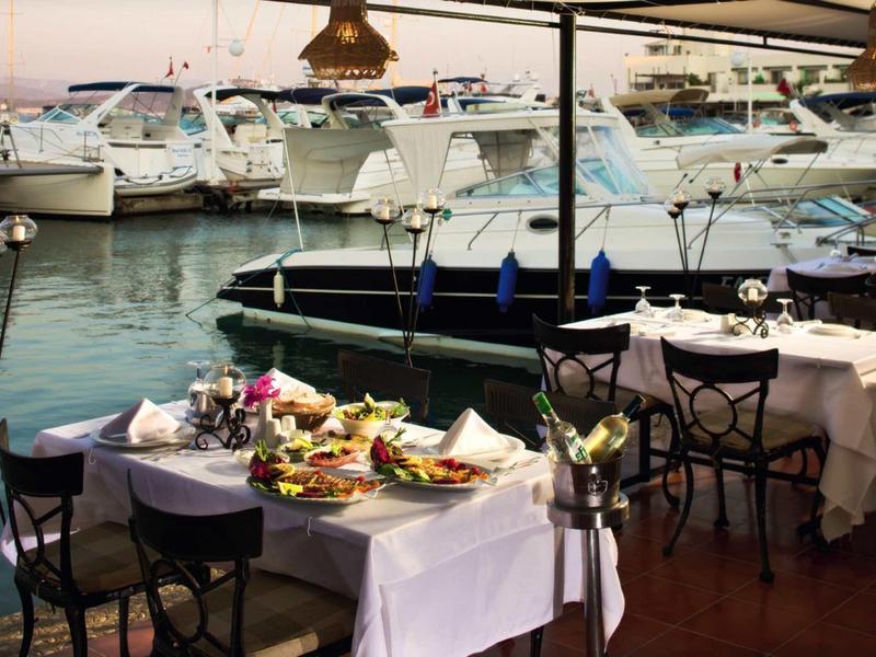 Restaurantterrasse mit weiß gedeckten Tischen am Hafen, dahinter liegen mehrere weiße Boote im Wasser.
