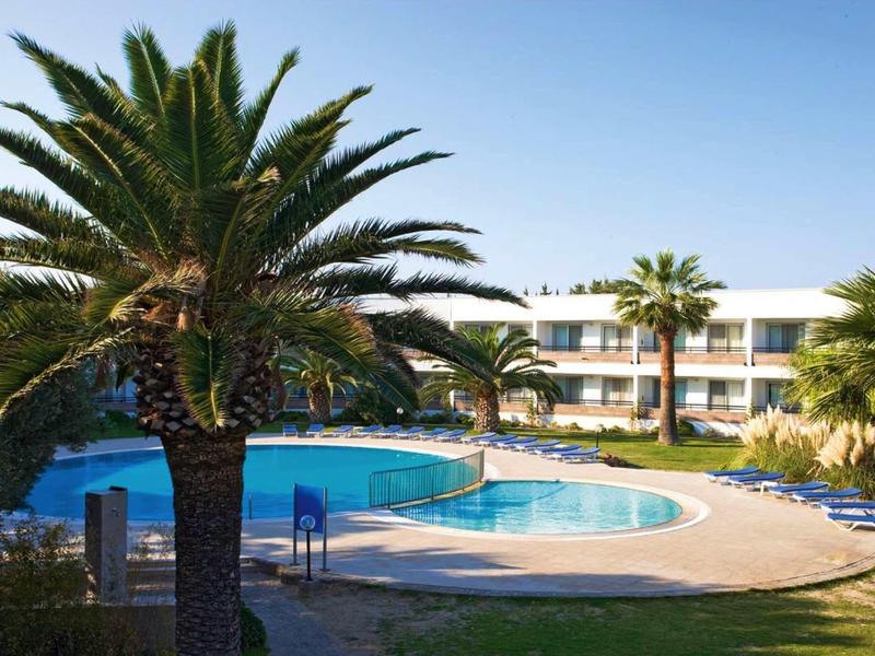 Ein Außenpool mit klar blauem Wasser, umgeben von Palmen und einem weißen Gebäude im Hintergrund.