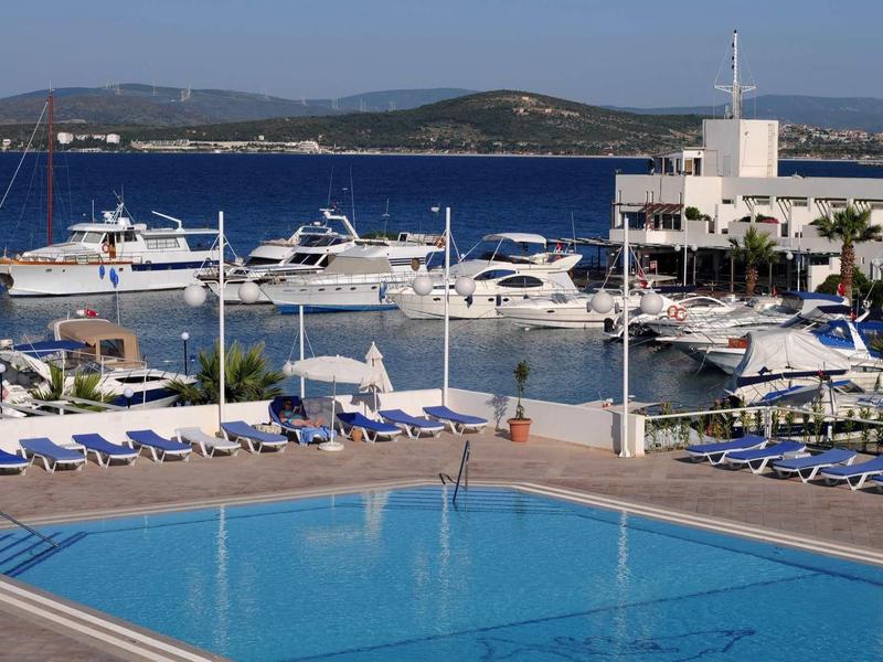 Blauer Pool mit Sonnenliegen am Hafen, dahinter weiße Yachten und sanfte Hügel im Hintergrund.