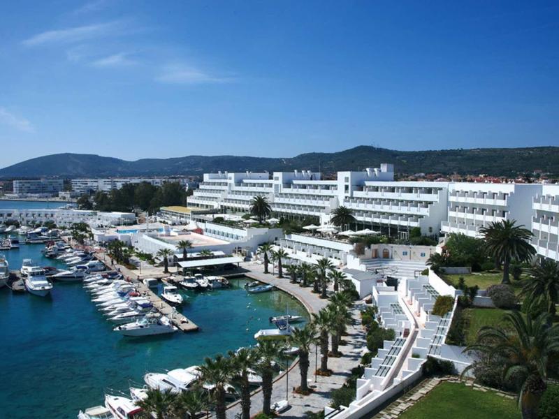 Großes weißes Hotel am Meer mit Yachthafen, Palmen und Bergen im Hintergrund unter blauem Himmel.