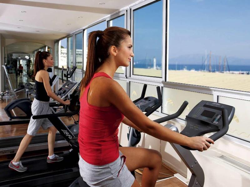 Zwei Frauen trainieren auf Laufband und Fahrrad-Ergometer mit Meerblick durch große Fenster.